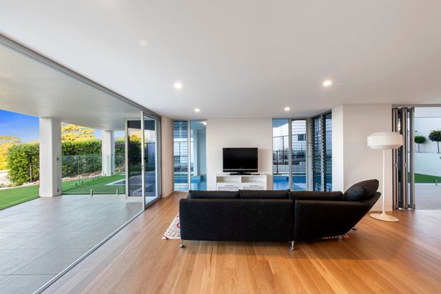 Bright, open-plan contemporary living room with warm timber flooring, a large dark sectional sofa, low white media console, and television. Full-height glazing and sliding glass doors connect the room to a covered outdoor terrace and landscaped garden, while recessed ceiling lights and a minimalist floor lamp complete the clean interior.