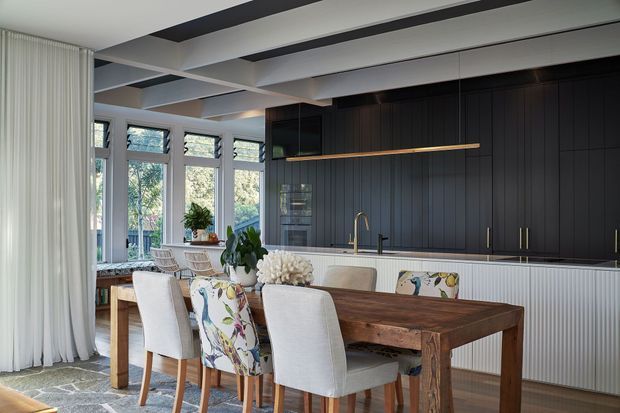A contemporary open-plan kitchen and dining area with a large rustic timber dining table surrounded by upholstered chairs. The kitchen features dark vertically panelled cabinetry, a long integrated timber shelf, a light ribbed island front, and a brass-toned faucet. Full-height windows and sheer curtains bring in abundant natural light, while exposed ceiling beams add architectural definition.