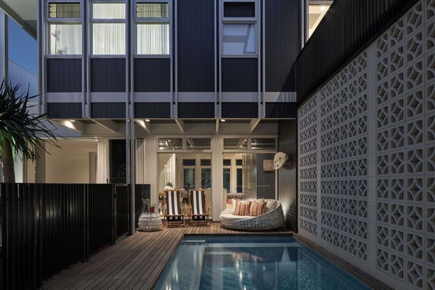 A compact contemporary courtyard viewed from an outdoor deck, featuring a narrow swimming pool, dark vertical metal cladding, large glazed doors and windows, timber decking, and a decorative concrete breeze-block wall. The two-storey composition includes an upper-level overhang and illuminated ground-floor entertaining area with outdoor lounge furniture.