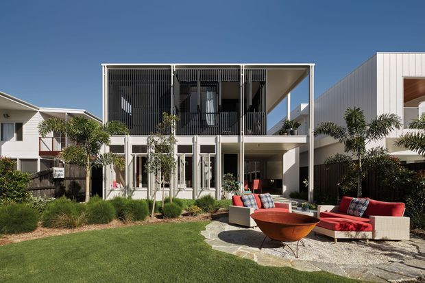 A bright contemporary two-storey residence viewed from a landscaped courtyard. The building has a rectilinear form, white rendered walls, extensive glazing, a deep upper-level balcony with dark vertical screening, and a flat roof. The courtyard includes lawn, ornamental planting, paved areas, and a red outdoor seating arrangement around a sculptural fire bowl.