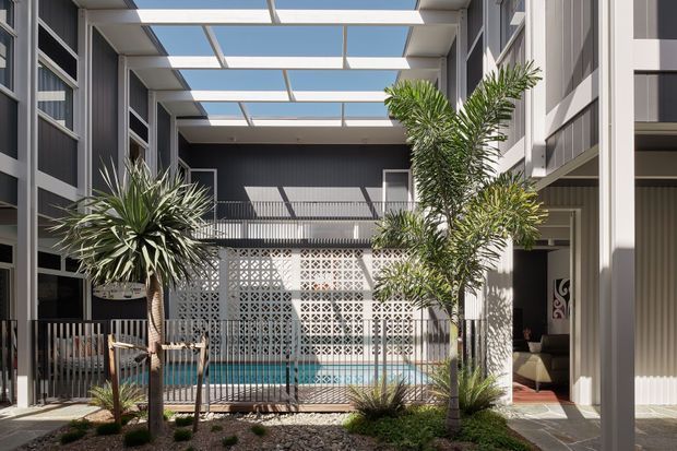A bright contemporary internal courtyard viewed from an outdoor walkway, enclosed by multi-storey building wings. The courtyard has a small swimming pool behind a metal safety fence, tropical planting, a patterned breeze-block privacy wall, and a translucent roof structure with repeated skylight panels. White and dark-grey architectural finishes create a crisp, high-contrast resort-like atmosphere.