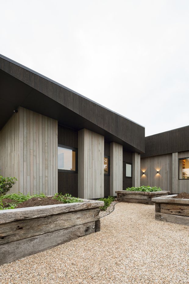 Exterior view of a contemporary single-storey building arranged around a gravel courtyard. The facade features dark vertical timber cladding, lighter vertical timber-clad projecting volumes, deep roof overhangs, rectangular windows, and warm wall-mounted lighting. Raised weathered timber planter boxes with low greenery line the courtyard.