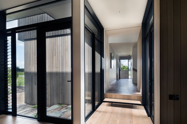 A contemporary residential hallway and entry zone with a long sightline toward an exterior opening. The space features pale timber flooring, white walls and ceiling, dark-framed full-height glazing and internal doors, and a raised transition with a timber step. Vertical timber cladding and a landscaped exterior are visible through the entry glazing.