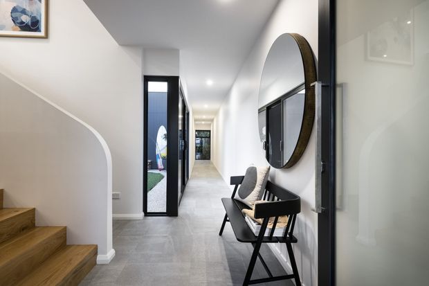 A bright contemporary residential hallway with a timber staircase, curved stair wall, pale grey tiled flooring, white walls, black-framed glazing and doors, and a black occasional chair beneath a large round wall mirror. The corridor extends toward a glazed exterior door and garden beyond.