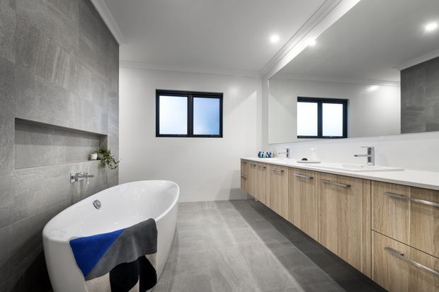 A spacious contemporary bathroom with a freestanding white bathtub, double vanity, large wall mirror, and wall-mounted fixtures. The room features grey large-format tiled walls and flooring, light timber-look vanity cabinetry, white benchtops, black-framed windows, and recessed ceiling lighting.