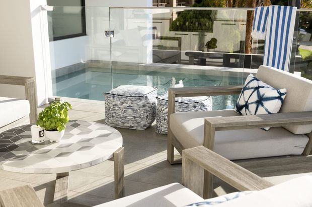 Bright outdoor terrace with a pale tiled floor, glass balustrade, and a small plunge pool or spa immediately beyond the seating area. The space is furnished with light timber-framed lounge chairs, white cushions, patterned ottomans, a round geometric coffee table, potted greenery, and a blue-and-white striped deck chair. The overall feel is airy, relaxed, and coastal.