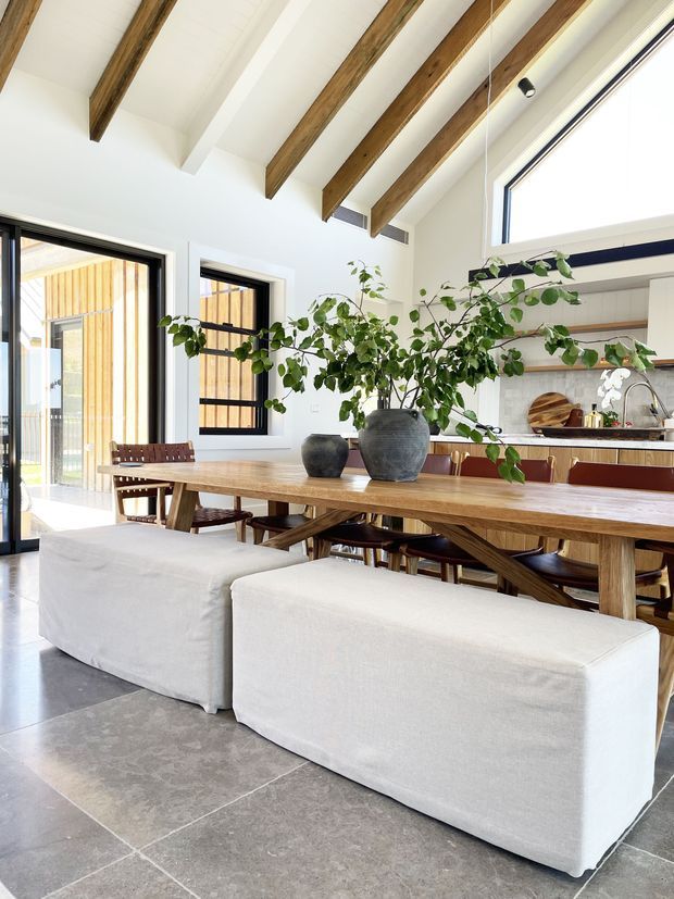 Bright open-plan dining and kitchen interior with a long solid timber dining table, timber chairs, white upholstered bench seating, large concrete-look floor tiles, and abundant natural light. The room features exposed timber ceiling beams, black-framed glazing, and a kitchen with white cabinetry, dark lower joinery, and a tiled splashback. Large ceramic vases with leafy branches form a central table arrangement.