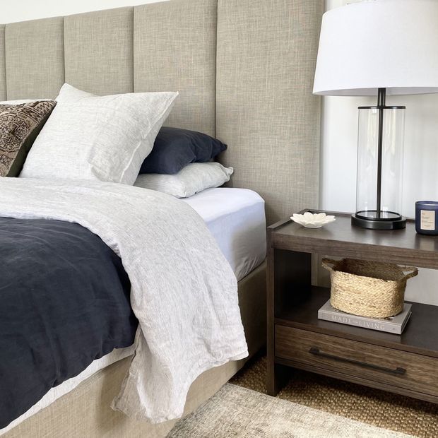 A softly styled bedroom featuring a large upholstered light-beige vertical-panel headboard, layered white and charcoal bedding, and a dark wood bedside table with an open shelf and drawer. A modern table lamp, woven basket, small decorative dish, candle, and neutral textured rug complete the warm, refined scheme.