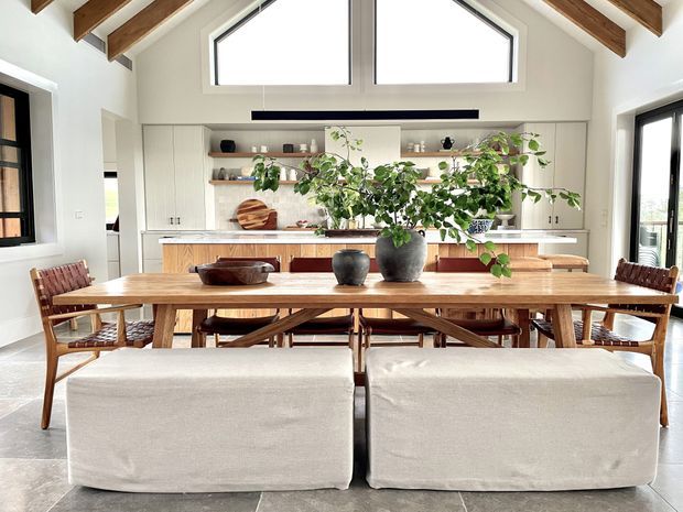 A bright open-plan dining and kitchen interior with a long solid timber dining table, woven timber dining chairs, upholstered bench seating, and large potted plants. The kitchen features light cabinetry, open timber shelving, a pale benchtop, and a timber island. Exposed timber ceiling beams and high clerestory-style glazing create an airy, warm contemporary setting.