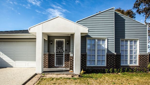 Front exterior view of a single-storey suburban home with a prominent gabled facade, pale horizontal weatherboard cladding, dark brick lower walls, white trim, multi-pane windows, a covered entry porch with decorative security door, and an attached double garage.