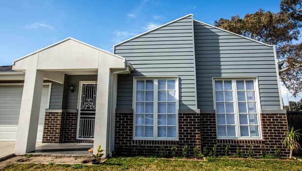 Front exterior view of a suburban single-storey home with a prominent gabled facade. The building combines dark horizontal weatherboard cladding, dark brickwork at the lower walls, rendered entry columns and gable detail, and white-framed multi-pane windows. A covered porch and security screen door define the entry, with a garage visible at left and modest front landscaping.