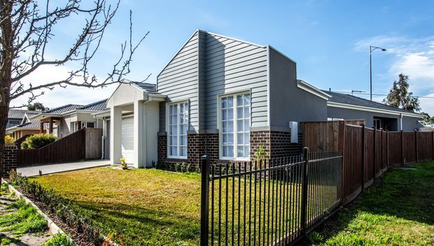 A contemporary suburban single-storey home viewed from the front, featuring a steeply articulated gable-like central facade clad in horizontal grey weatherboards, dark brick at the lower wall, white-framed windows, a dark tiled roof, timber fencing and a small front lawn.