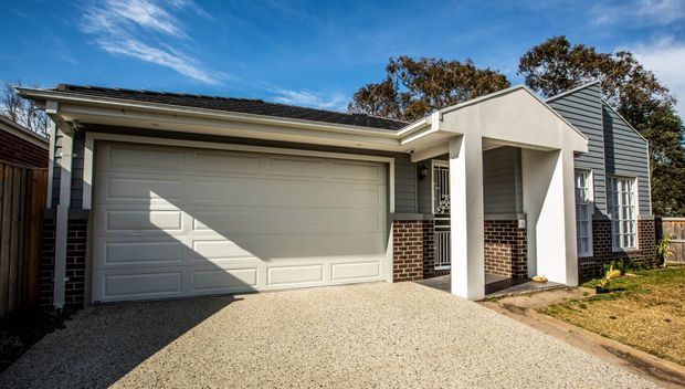 Street-facing exterior of a compact suburban home with a wide double garage, pale rendered entry porch, grey horizontal cladding, brickwork accents, gabled roof sections, and a textured driveway. Bright daylight highlights the clean contemporary facade.