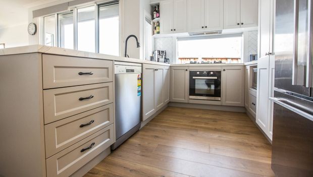Bright, compact U-shaped kitchen with a central island, white shaker-style cabinetry, brushed metal appliances, a timber-look floor, and a pale patterned tile splashback. Large windows provide abundant natural light.