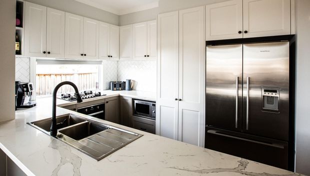 Bright, traditionally detailed white kitchen with extensive shaker-style cabinetry, a large marble-look island with undermount sink and gooseneck tap, stainless-steel French-door refrigerator, integrated cooking appliances, and a white geometric tiled splashback. Natural light enters through the window above the cooktop.