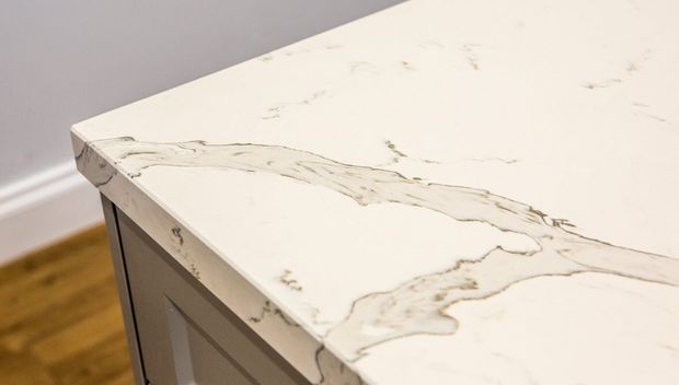 Close-up of a white stone kitchen benchtop with prominent grey veining and a thick eased edge, set above light painted cabinetry. A timber floor and neutral wall are visible in the background.