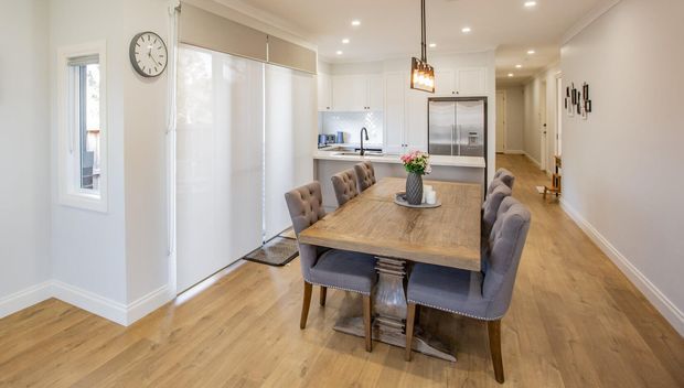 Bright open-plan dining area with a timber dining table and upholstered chairs, connected to a contemporary white kitchen. The room features warm wood-look flooring, white cabinetry, roller blinds, recessed ceiling lights, and a pendant light above the table.