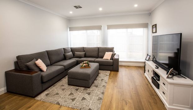 Bright contemporary living room with a large dark grey L-shaped sectional sofa, matching ottoman, patterned area rug, white media console and wall-mounted television. The room has pale walls, recessed ceiling lights, wide windows with roller blinds, and warm timber flooring.