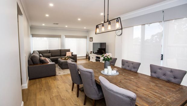 Open-plan living and dining area with warm timber flooring, a large rustic timber dining table and upholstered dining chairs in the foreground, and a dark sectional sofa with television at the rear. Neutral walls, grey Roman blinds, recessed ceiling lights, and a linear black pendant fixture create a contemporary, comfortable interior.