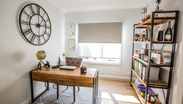 A bright, compact home office with a rustic timber desk, upholstered chair, open black metal-and-wood shelving filled with books and decor, a large industrial-style wall clock, and a roller blind over the window. The room has light walls, timber flooring, and a refined eclectic feel.