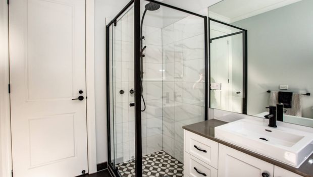 Bright contemporary bathroom featuring a frameless glass shower enclosure with black metal framing, white marble-look wall tiles, a patterned black-and-white floor, and a white vanity with dark benchtop and black tapware. A large mirror reflects the vanity and shower area.