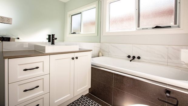Bright, compact bathroom with pale green walls, white framed windows, a white vanity with shaker-style cabinetry and dark hardware, a white basin and black tapware, and a built-in bathtub. The bath is clad in dark brown tiled panels with a white tiled splashback featuring subtle veining. A patterned black-and-white floor adds contrast.