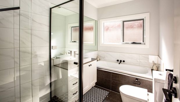 Bright contemporary bathroom featuring a glass-enclosed shower, freestanding-style white vanity with mirrored cabinet, built-in bathtub, wall-mounted toilet, and large frosted windows. White marble-look wall tiles contrast with dark charcoal bath paneling and patterned floor tiles.