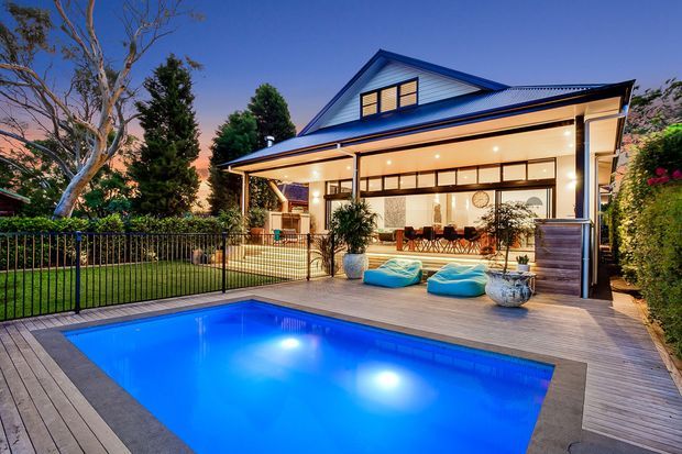 A dusk exterior view of a two-storey gabled home with a broad covered outdoor entertaining area, illuminated interior glazing, timber decking, landscaped garden, and a bright blue swimming pool in the foreground. The house features pale horizontal cladding, dark blue roof trim, and a contemporary poolside lounge setting.