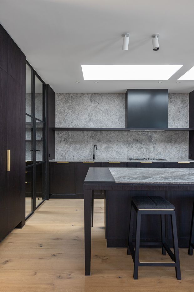 A sophisticated contemporary kitchen with dark timber cabinetry, black-framed glazed doors, a grey stone backsplash and benchtop, integrated cooktop and rangehood, and a central island with bar seating. Pale timber flooring and a large ceiling skylight balance the dark joinery, while brass-toned hardware adds a subtle accent.