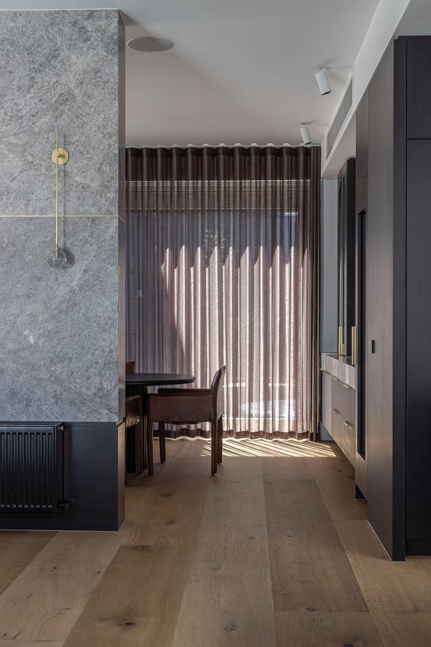 A refined contemporary interior showing a compact dining area beyond a stone-clad partition. The space features wide-plank light timber flooring, dark built-in cabinetry with brass-toned handles, a dark dining table and chairs, sheer brown curtains filtering strong daylight, a black radiator, recessed and directional ceiling lighting, and an integrated linear air-conditioning vent.