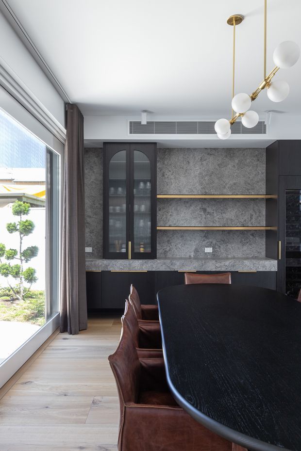 A refined dining room with a large black oval dining table and dark brown leather dining chairs. The room features dark built-in cabinetry with fluted-glass doors, open timber shelves, a textured grey stone-look wall finish, pale timber flooring, full-height glazing with curtains, and a sculptural brass-and-glass pendant light. A landscaped garden is visible outside.