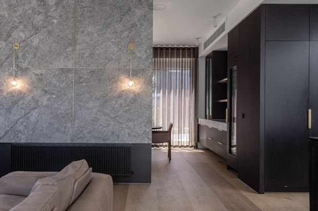 Contemporary open-plan living area with a large grey stone feature wall, pendant lighting, a low dark radiator, and a pale upholstered sofa in the foreground. The room extends toward a dining area with sheer curtains and is lined with tall dark timber cabinetry and integrated shelving, creating a refined, moody interior.