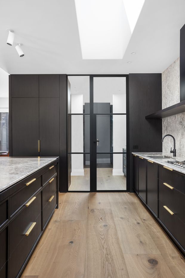 A refined contemporary kitchen with dark timber cabinetry, brass pulls, pale natural timber flooring, and veined stone benchtops. A black-framed glazed door separates the kitchen from a bright hallway, while a large skylight and recessed lighting provide abundant illumination. The right wall features a textured pale stone or tile splashback and open dark shelving.