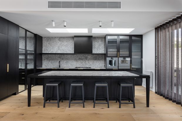 A sophisticated contemporary kitchen with a large dark island and four black bar stools, matte black cabinetry, grey stone splashback, integrated appliances, and light timber flooring. Floor-to-ceiling glazed doors and sheer curtains line the right side, while skylights and recessed spotlights provide natural and artificial illumination.