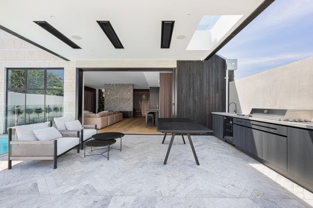 A bright contemporary outdoor entertaining area with a covered patio, pale stone herringbone flooring, built-in dark cabinetry and a long outdoor kitchen with sink and barbecue. A dark timber-clad wall and broad opening connect the patio to an interior living room, while an outdoor lounge setting occupies the foreground. Linear recessed ceiling lights and a large skylight add to the modern architectural character.