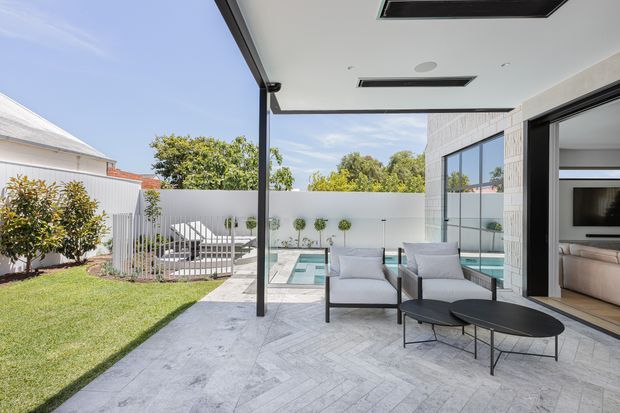 A bright contemporary outdoor entertaining area with a covered patio opening onto a swimming pool and landscaped backyard. The space features pale herringbone-pattern paving, black-framed full-height glazing and sliding doors, a white rendered boundary wall, and minimalist outdoor lounge furniture. A living room is visible through the open glazing.