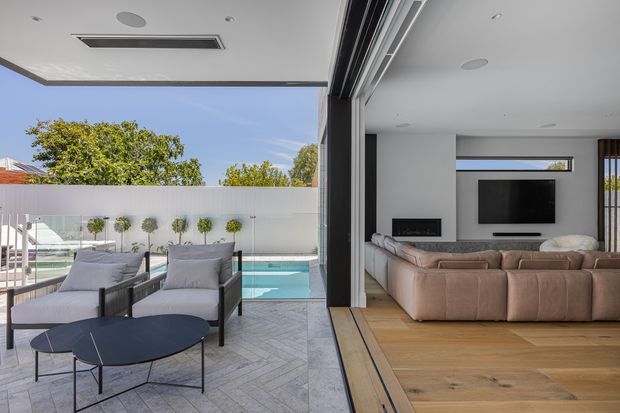 Contemporary indoor-outdoor living area with large retractable glazing opening onto a tiled patio and swimming pool. The interior features a neutral sectional sofa, wall-mounted television, built-in fireplace, timber flooring and recessed ceiling lighting, while the outdoor zone includes lounge chairs and a low round table.
