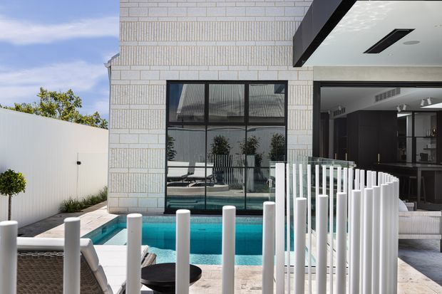 Contemporary residential pool courtyard viewed from an outdoor entertaining area, featuring a turquoise swimming pool, white vertical balustrade elements, pale masonry walls, large dark-framed glazing, and an open-plan interior beyond. The scene is bright and refined with strong geometric lines and indoor-outdoor connection.