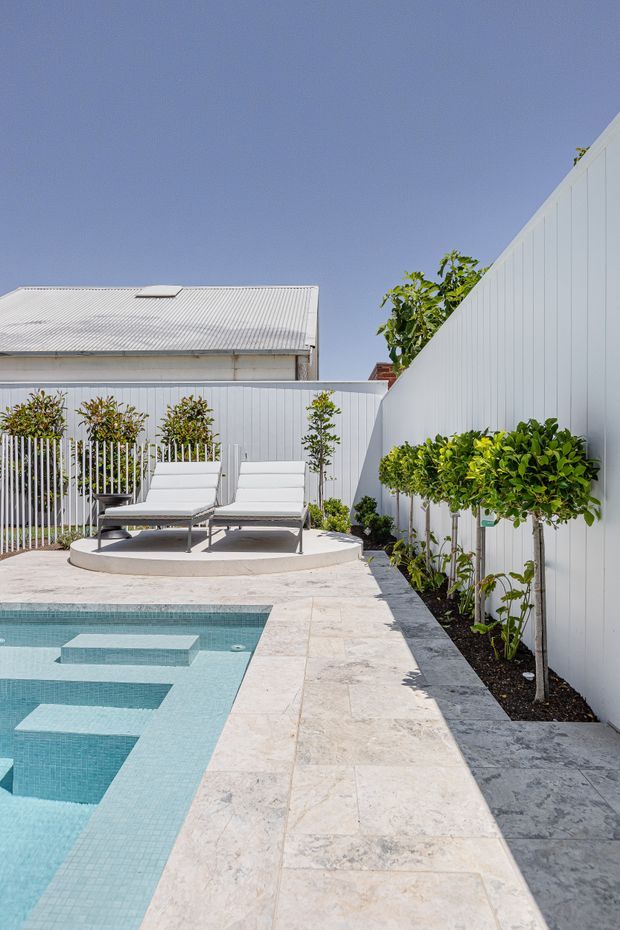 A bright, contemporary backyard pool area with pale stone paving, a turquoise tiled swimming pool, white vertical-panel fencing, and a raised circular lounging platform with two white chaise lounges. Slim-trunked trees and clipped greenery line the perimeter, creating a clean, private courtyard-like outdoor setting.