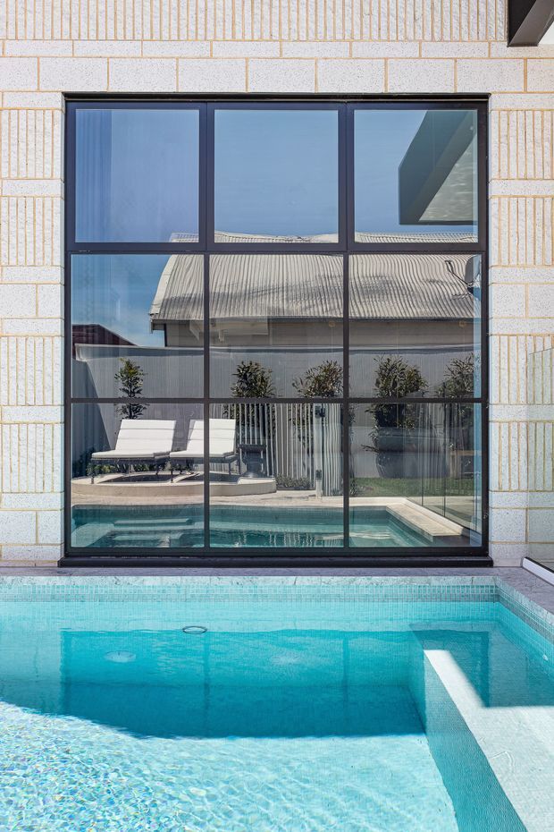 A close exterior view of a large black-framed grid window set into a pale textured masonry facade, looking onto a bright turquoise swimming pool. The glazing reflects a neighbouring corrugated roof and courtyard planting, while poolside loungers and a landscaped courtyard are visible beyond the window. The scene is crisp, contemporary, and strongly geometric.
