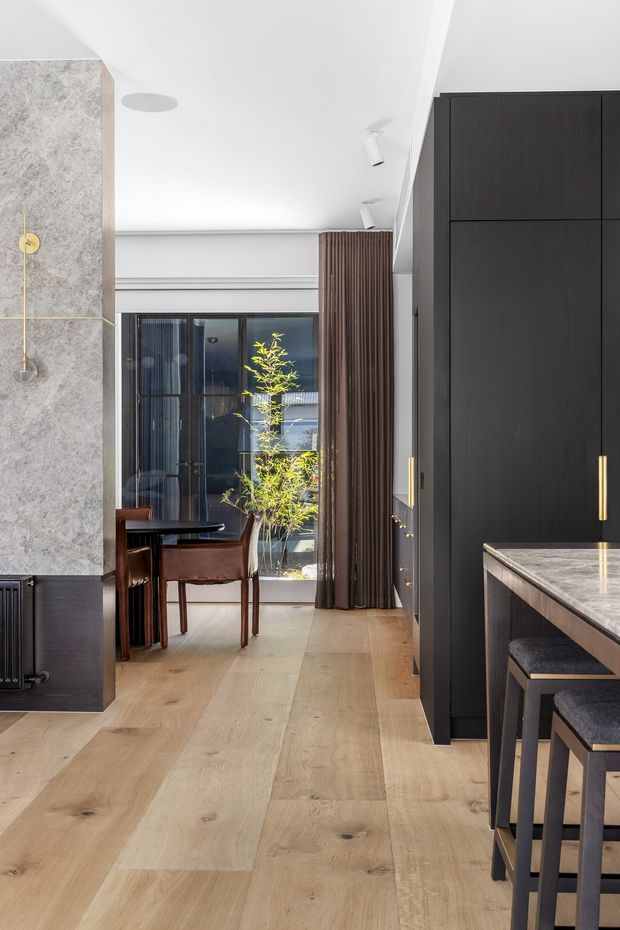 A refined open-plan interior showing a kitchen in the foreground and a dining area beyond. The space features wide-plank light timber flooring, dark charcoal built-in cabinetry with brass-toned handles, a pale stone kitchen benchtop, a textured stone-clad fireplace or partition, full-height dark curtains, and a black-framed glazed opening. A round dining table and leather dining chairs sit near the window, while stools line the kitchen island. The palette is contemporary, warm, and high-contrast.