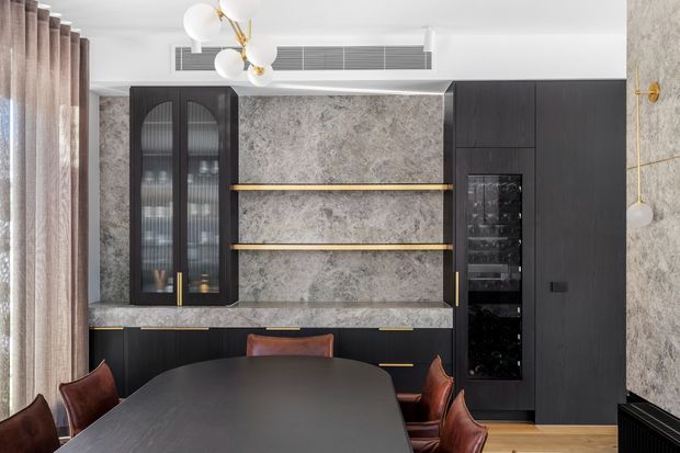 A refined dining area with a large dark dining table and leather chairs, framed by a full-height dark built-in cabinetry wall. The joinery incorporates glass-front display cabinets, open shelving with brass or gold trim, a stone-look feature surface, and an integrated wine or beverage cabinet. Soft curtains, sculptural globe pendant lighting, timber flooring, and a marble-patterned wall create a luxurious contemporary setting.