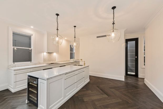 Bright, refined kitchen with a large white paneled island, integrated wine cooler, pale stone benchtops, white cabinetry, pendant lighting, and dark herringbone timber flooring. The room has decorative wall and ceiling mouldings, tall windows, and a glass-panelled door.