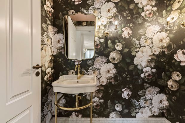 A compact powder room with a wall-mounted white basin on a gold metal stand, exposed gold plumbing, a rounded rectangular mirror, a white paneled door, and dramatic dark floral wallpaper covering the walls. The room has a refined, vintage-inspired decorative character.