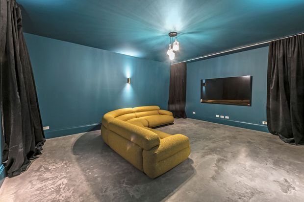 A moody home cinema room with deep teal walls and ceiling, full-height dark curtains, a large mustard-yellow sectional sofa, a wall-mounted screen or television, dramatic wall sconces, and a polished concrete-look floor. The room has a compact, intimate layout with a dark, cinematic atmosphere.