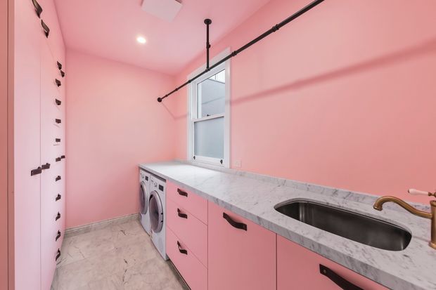 A compact, brightly styled laundry room with vivid pink walls and matching pink built-in cabinetry. The room features a long marble-look benchtop with an inset stainless-steel sink and brass-toned tapware, a front-loading washing machine, black drawer and cabinet handles, pale stone-look flooring, a window, and exposed black ceiling-mounted hanging rails.
