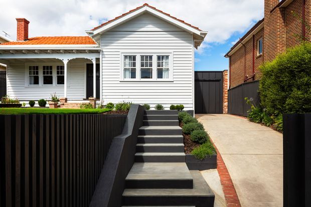 Street-facing view of a renovated single-storey house with a white weatherboard gabled facade, red tiled roof, front porch and windows. A landscaped front garden, dark vertical fence, concrete stairway and driveway sit between the house and adjacent brick building.