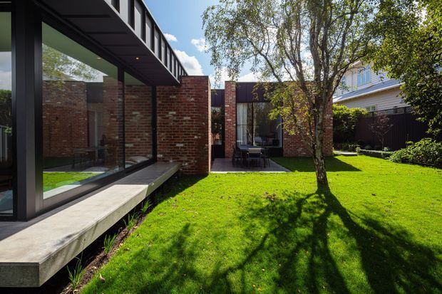 A contemporary single-storey residential extension arranged around a landscaped lawn and mature tree. The building combines dark-framed floor-to-ceiling glazing, exposed red brick walls, a projecting concrete ledge, and a dark metal roof or fascia. A small paved outdoor dining area is visible beyond the lawn, with a traditional neighbouring house in the background.
