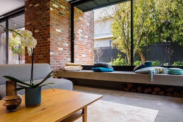 A bright contemporary living room with exposed red brick walls, expansive black-framed glazing, and a built-in window seat overlooking a leafy garden. The room includes a light timber coffee table, upholstered lounge seating, cushions, an orchid, and a timber-lined bench with stored firewood beneath.