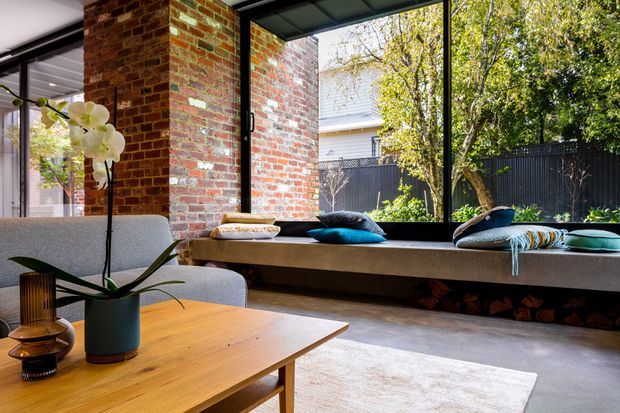 A warm contemporary living area with exposed red-brick walls, expansive black-framed glazing, and a long built-in window seat overlooking a leafy garden. A timber coffee table, upholstered armchairs, cushions, and an orchid create a relaxed, refined interior. The polished concrete floor and industrial-style glazing complement the brickwork.